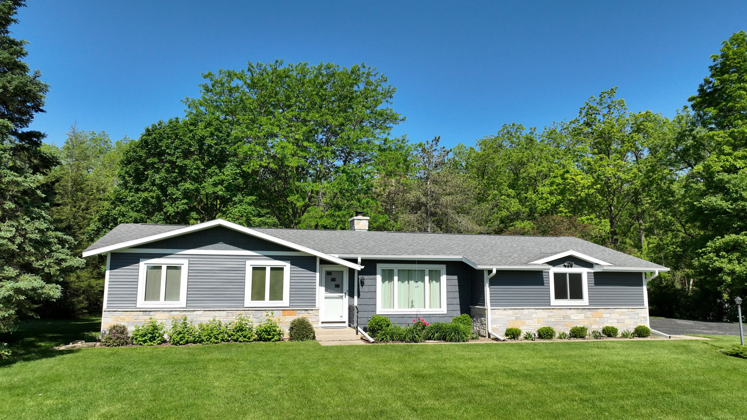 Single-story gray house with white trim, stone accents, a front door, large windows, and a green lawn with trees in the background under a clear blue sky.
